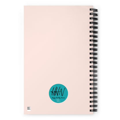A tannish pink background on the back of a spiral notebook with a teal colored logo with the letters HHN, the words Happy Healthy Nurses and a stethoscope in the shape of a heart within the teal circle logo